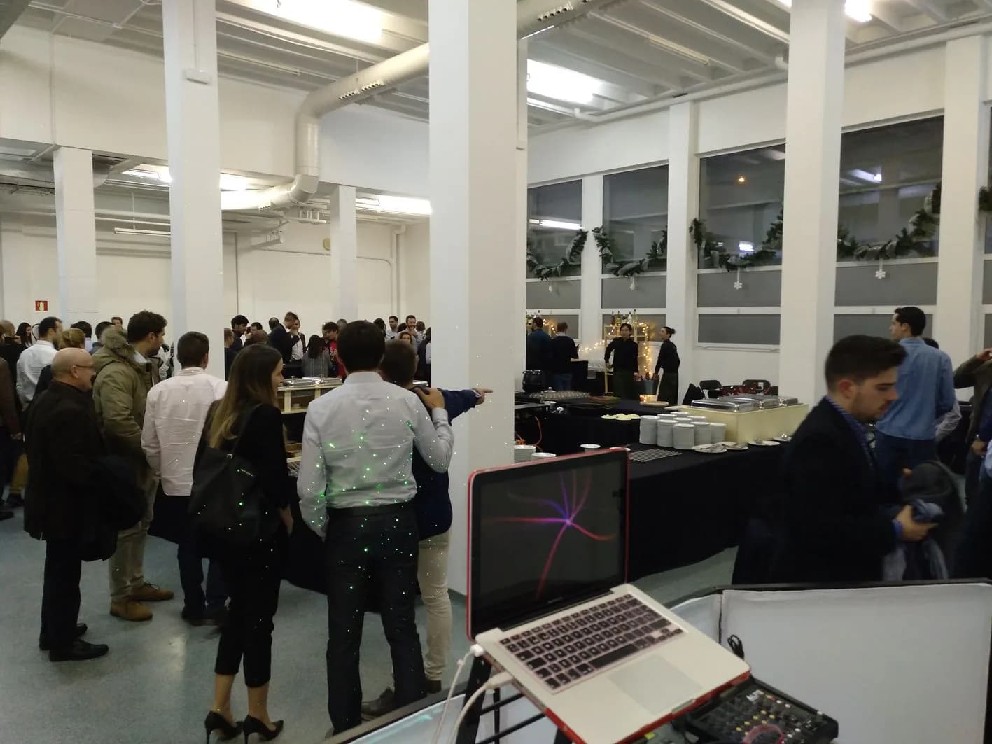 Cóctel de empresa con invitados conversando junto a buffet y MacBook con mesa de mezclas en sala industrial blanca