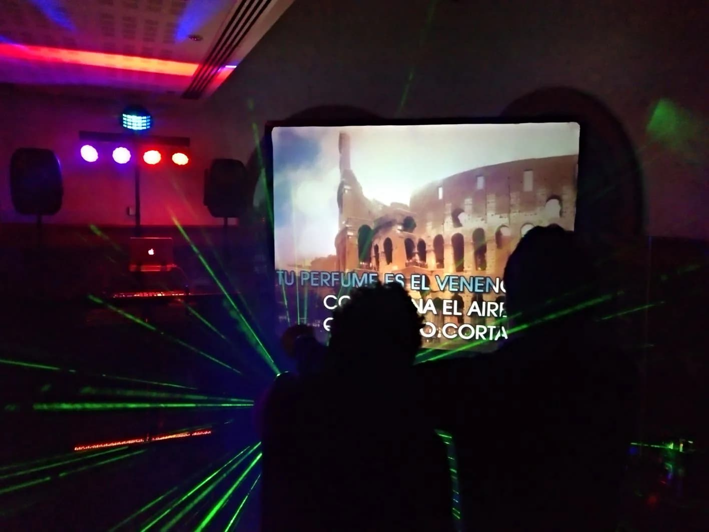 Sala oscura con pantalla de karaoke con letra de canción romántica, mesa de DJ al fondo y láseres verdes recorriendo la sala